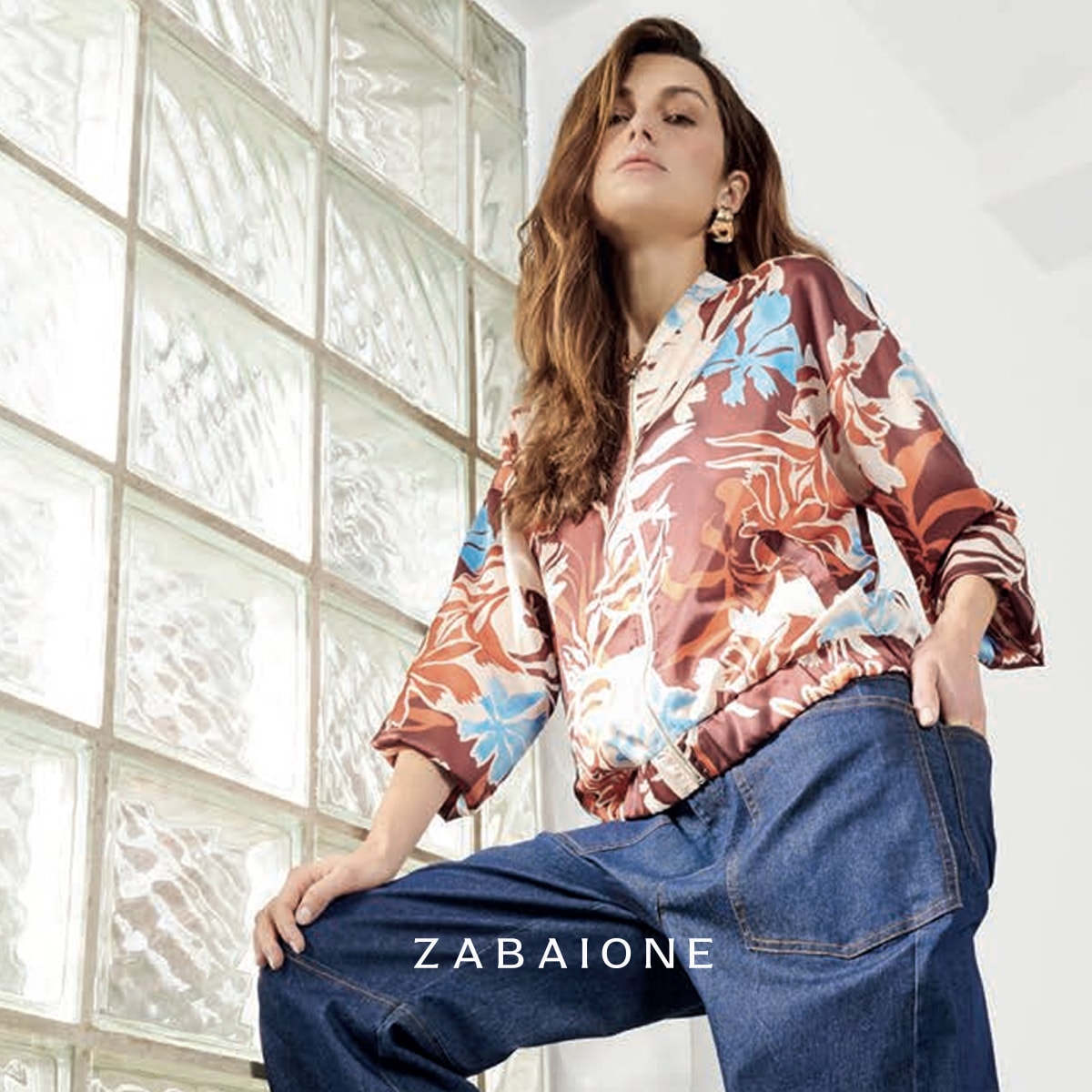 Young woman posing in a colorful floral blouse and blue denim pants against a textured glass block wall.